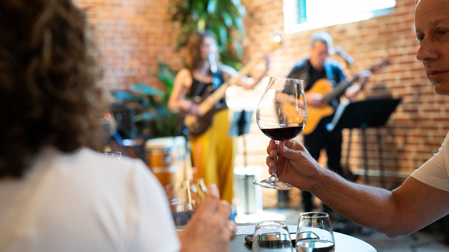Two musicians play on a small indoor stage with people drinking wine in the foreground.