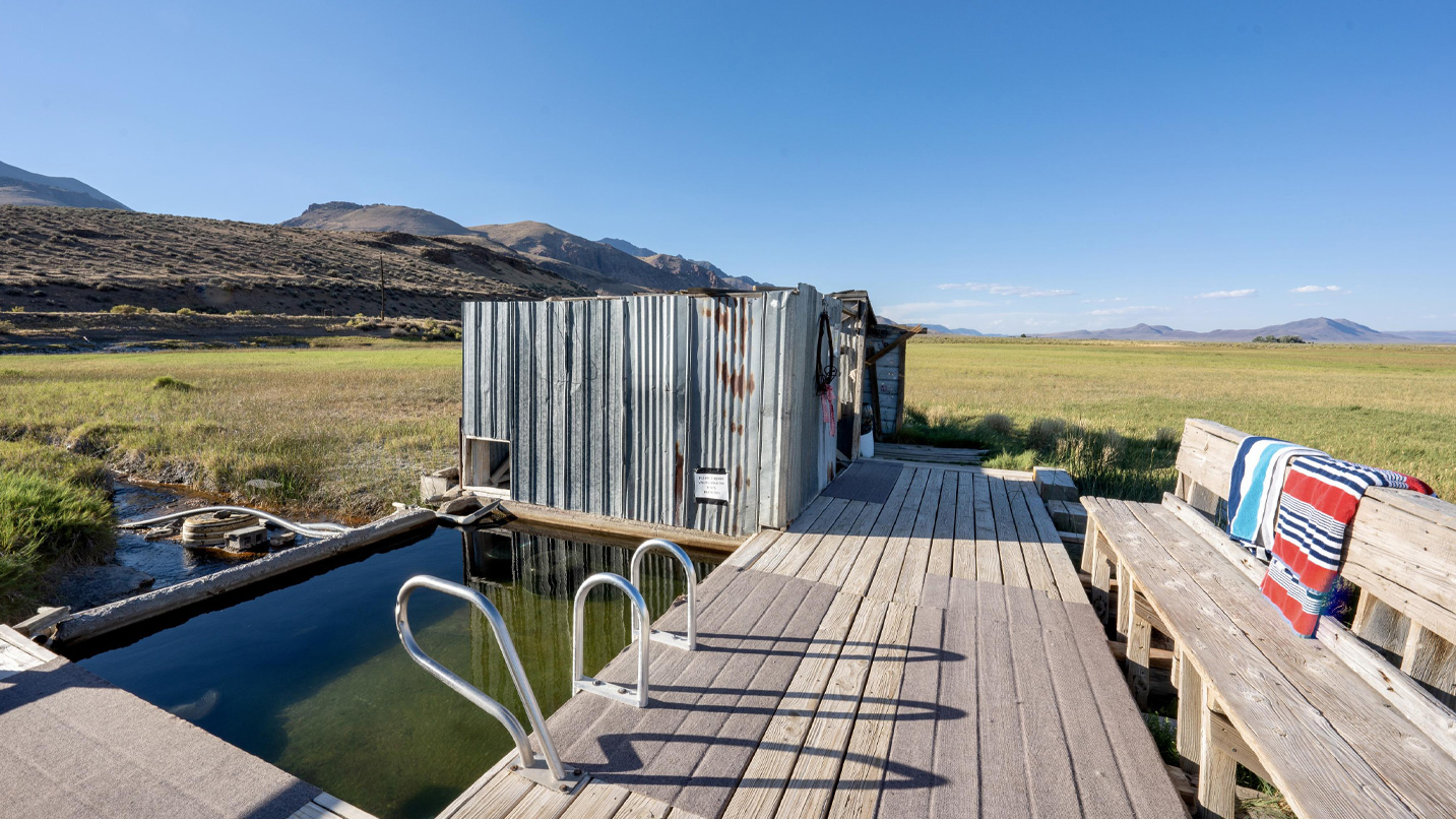 A small soaking pool is accessible by a short wooden planked area, with metal changing rooms and a wooden bench where towels hang.