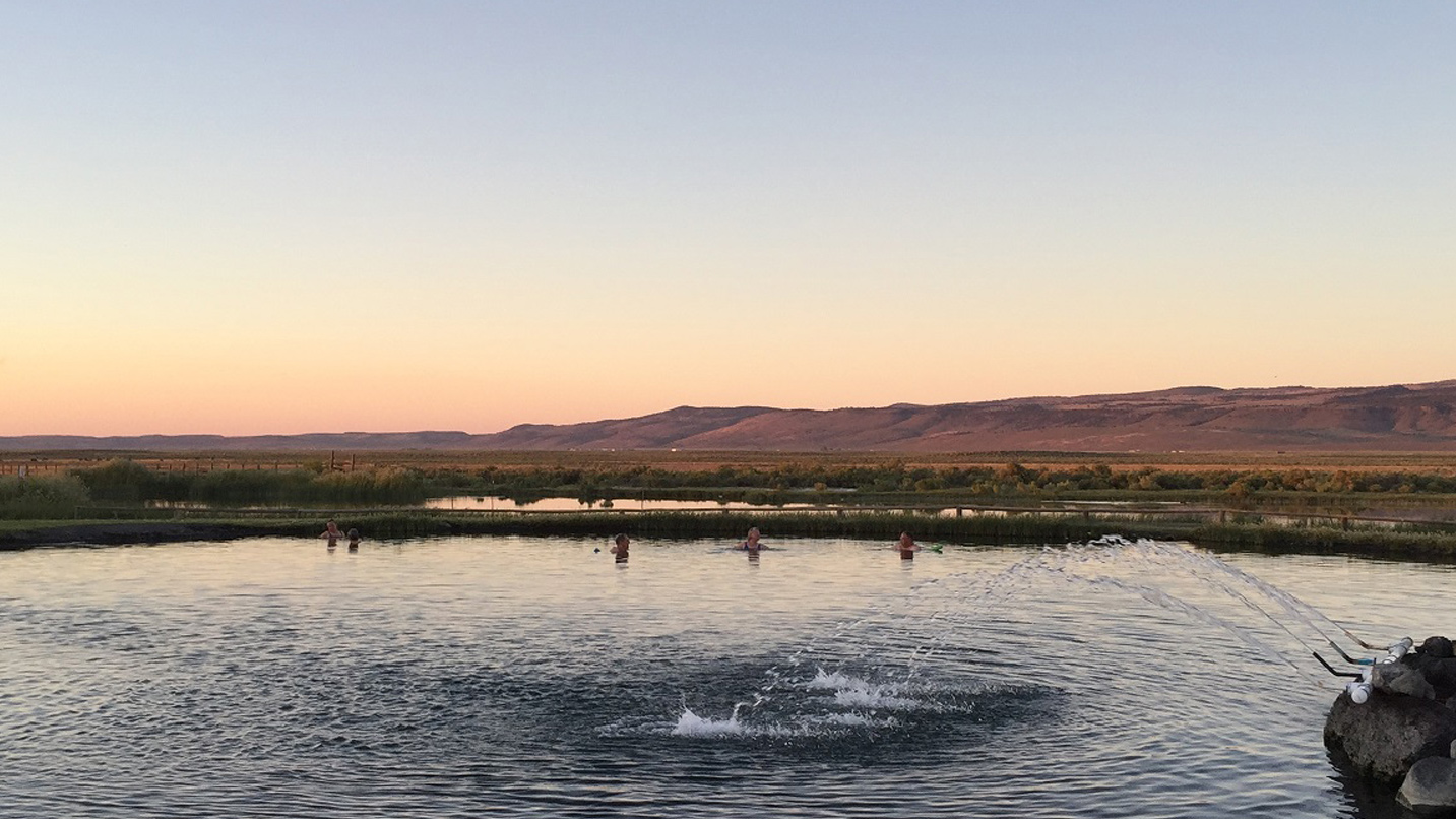 People soaking in a large hot spring against a high desert backdrop around sunset.