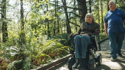 A woman in a power wheelchair and a man walking go along a boardwalk trail in a forested area.