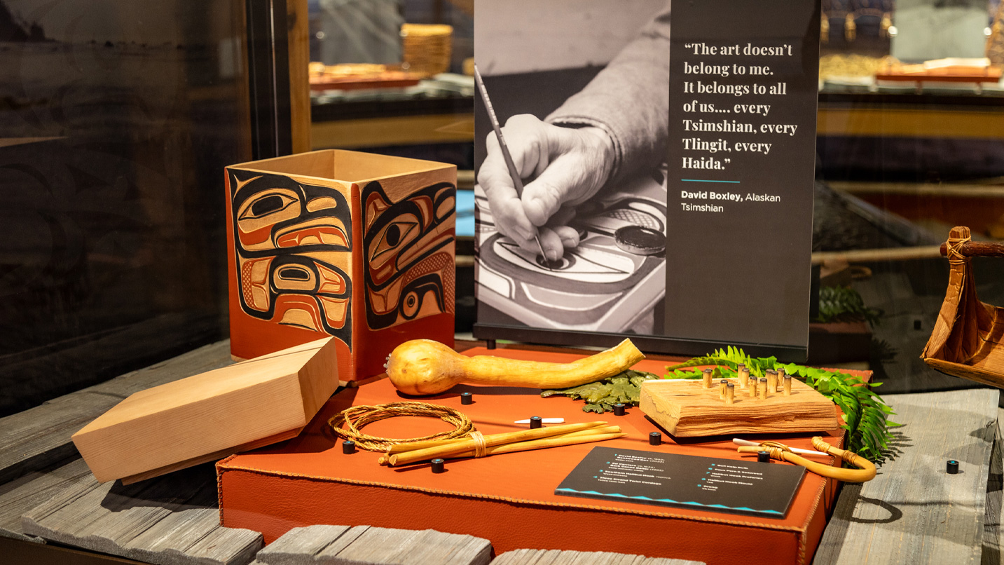 A wooden box with Indigenous designs is displayed alongside carving tools, a gourd, and leafy fern. Text reads: “The art doesn’t belong to me. It belongs to all of us... every Tsimshian, every Tlingit, every Haida." - David Boxley, Alaskan Tsimshian.