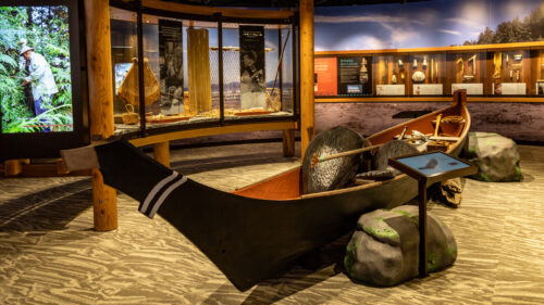A museum exhibit showcasing a traditional wooden canoe with informational displays and a video screen in the background.