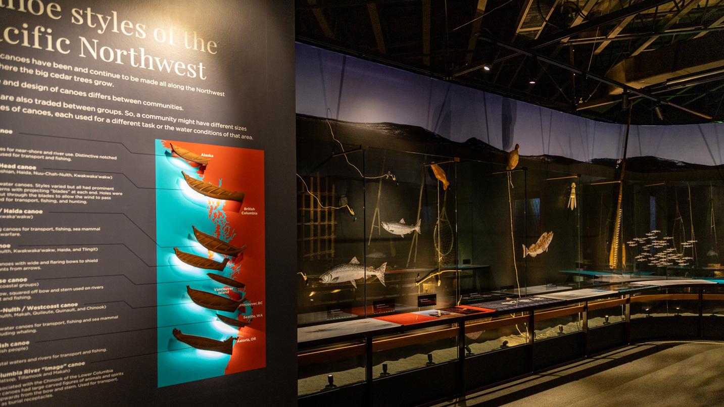 Exhibit featuring models of Pacific Northwest canoes illuminated on a wall; text reads "Canoe styles of the Pacific Northwest." Nearby, fish and fishing tools are displayed in glass cases.