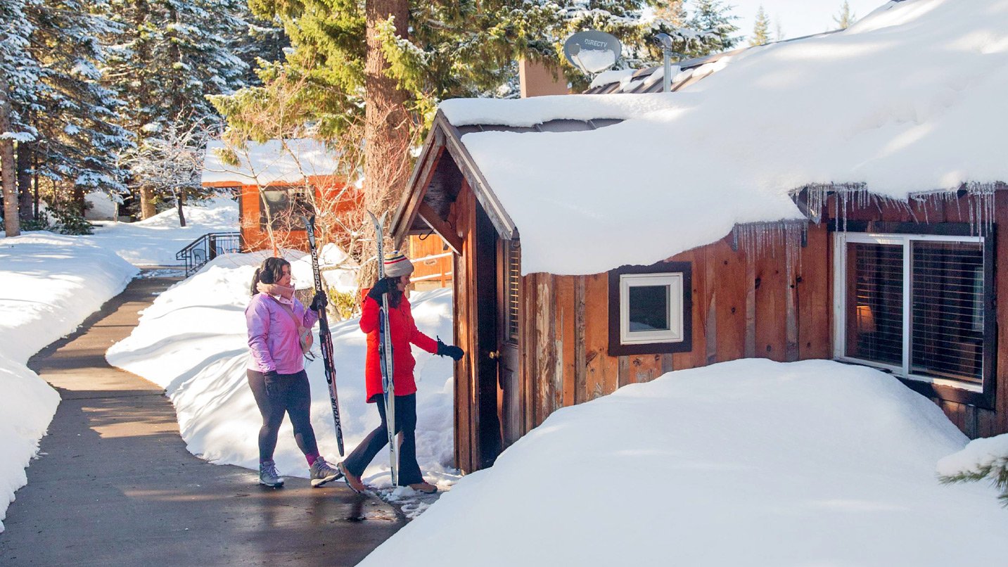 Two people carrying skis walk into a snowy cabin.