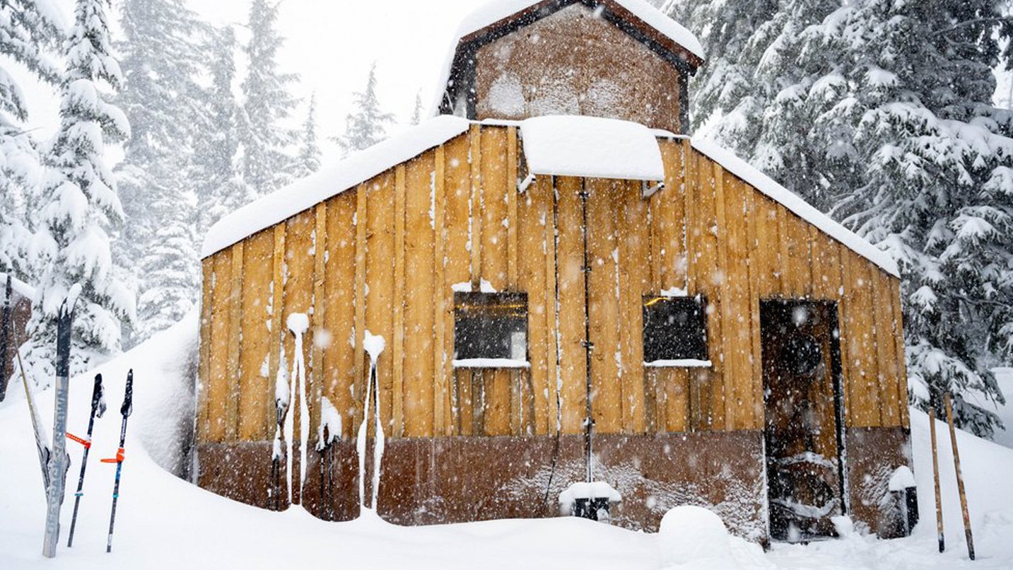 Skis planted in the snow in front of a wooden hut.
