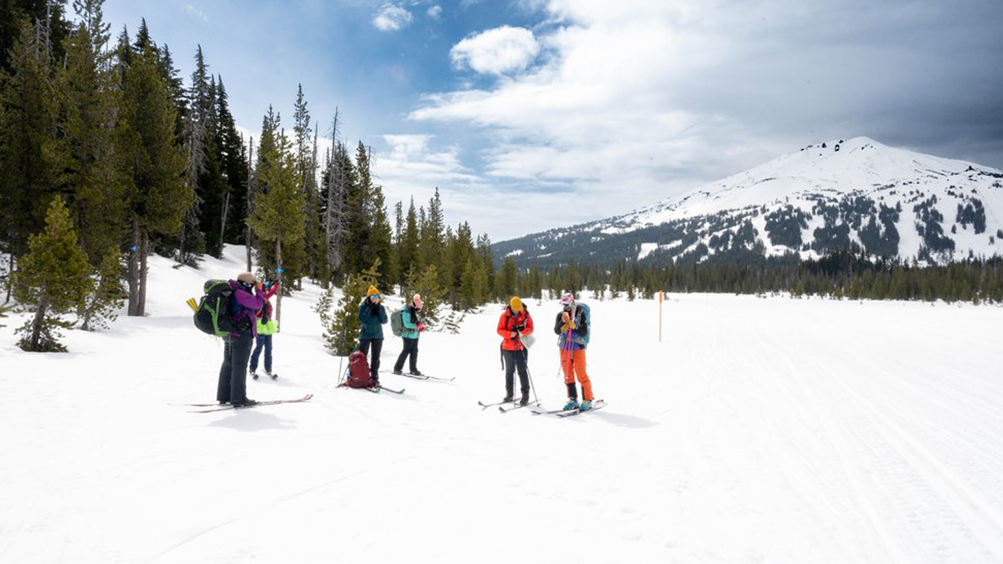 A group of people on skis gather around in the snow at the foot of a mountain.