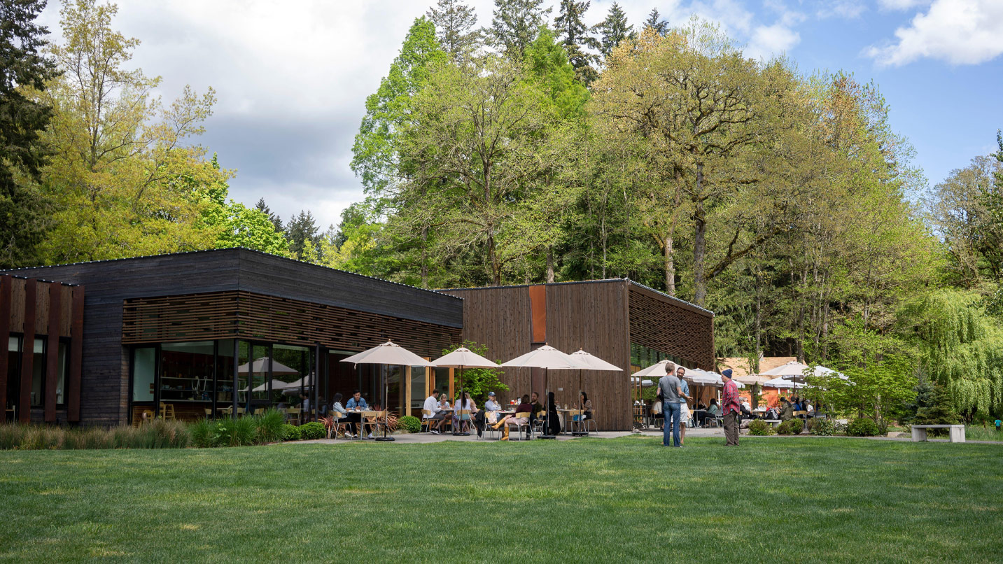 Modern building with green lawn and people standing and sitting on patio with umbrellas