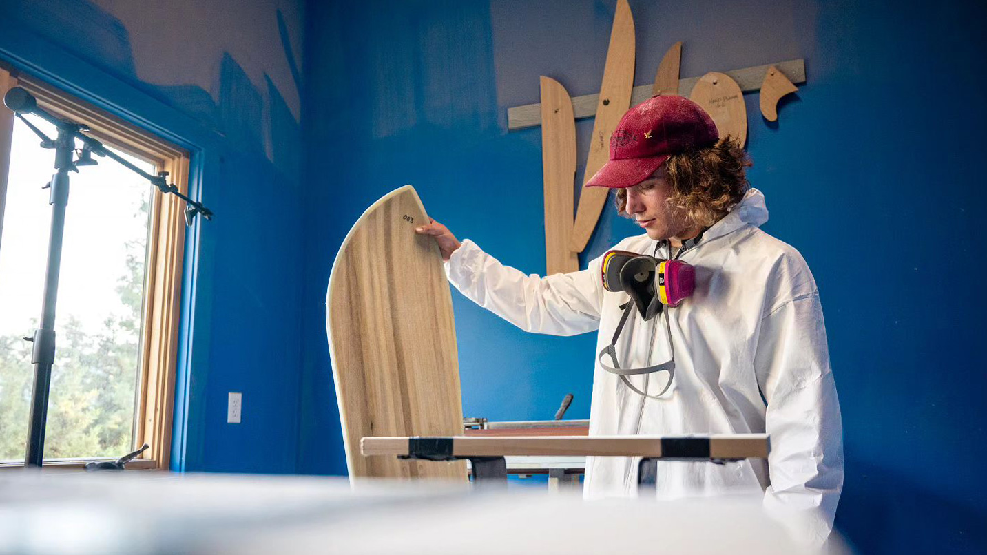 A young man in a work studio holds up a wooden snowboard.