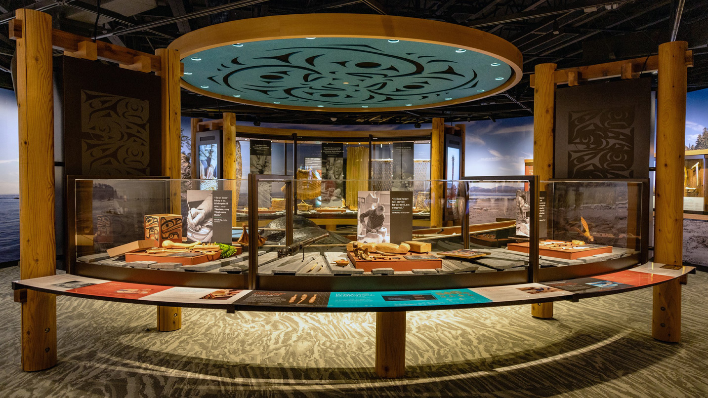 Exhibit with artifacts and informational displays is encased in glass on a circular platform. Wooden pillars and an intricately patterned ceiling create a cultural atmosphere. Text includes: "The air doesn't belong to us, 'Hida gwáay," and "Half of our story never gets told."