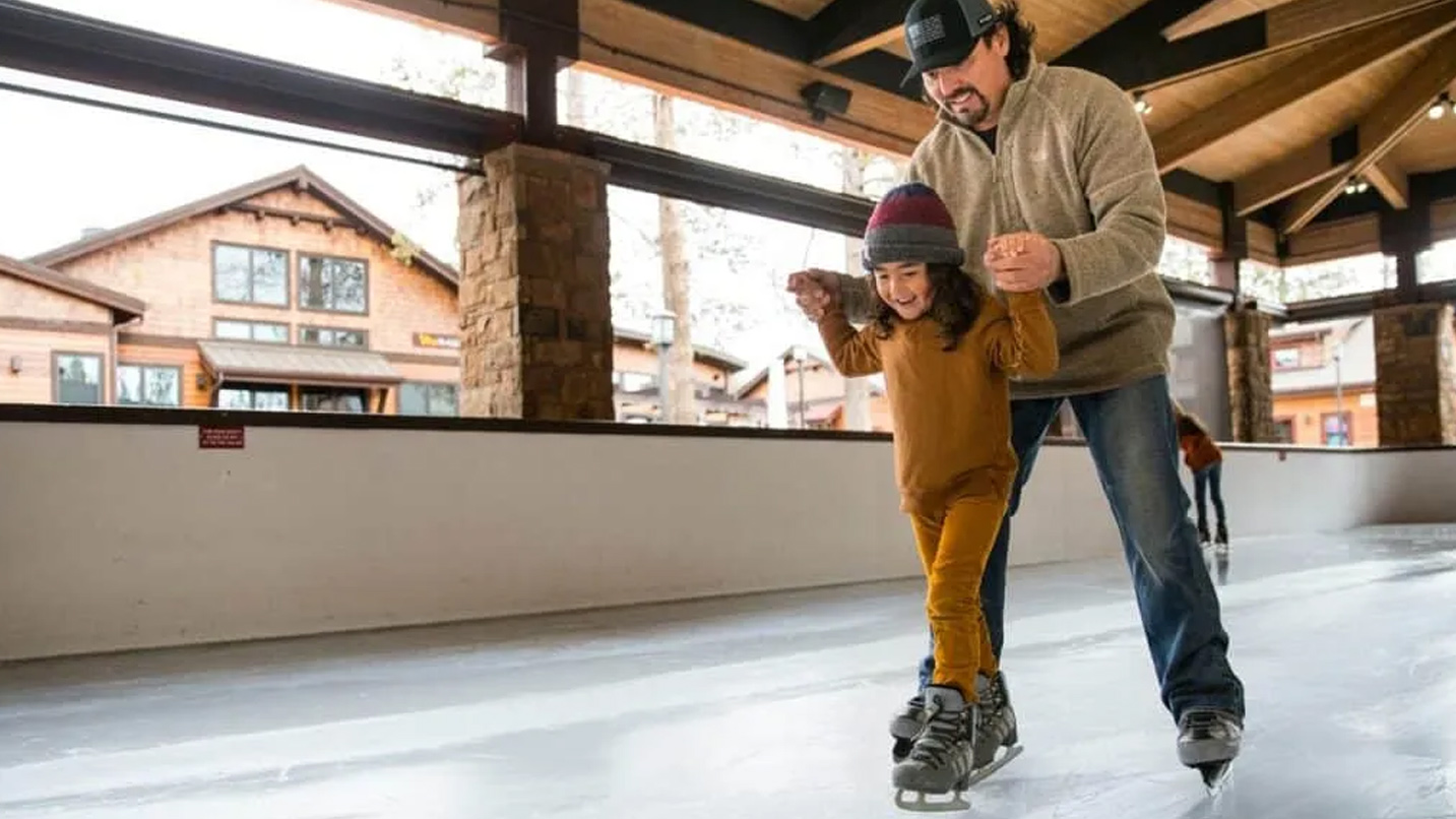 A man helps a young girl balance on ice skates on an ice rink.