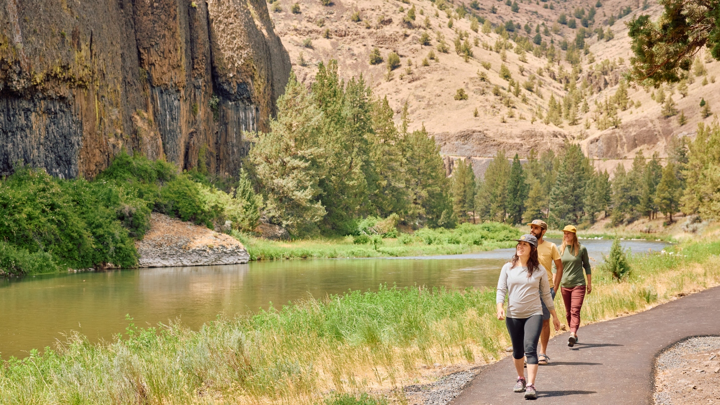 Three people walk on paved path next to grassy river canyon