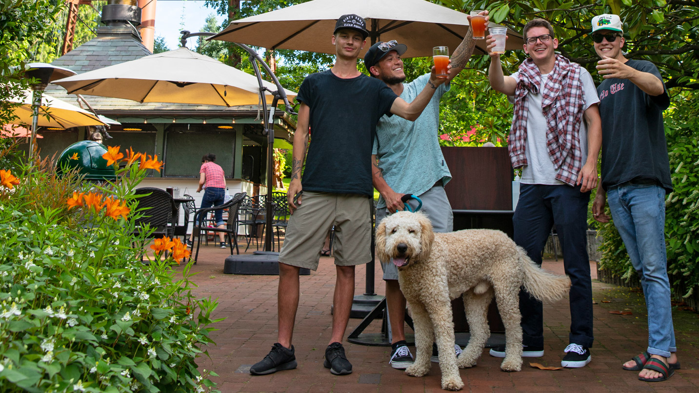 Four male friends cheer with brews at an outdoor seating patio. A labradoodle dog also smiles at the camera.