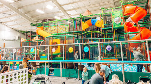 A large indoor play structure with winding slides, a ball pit and climbing structures. Several families watch children at play.