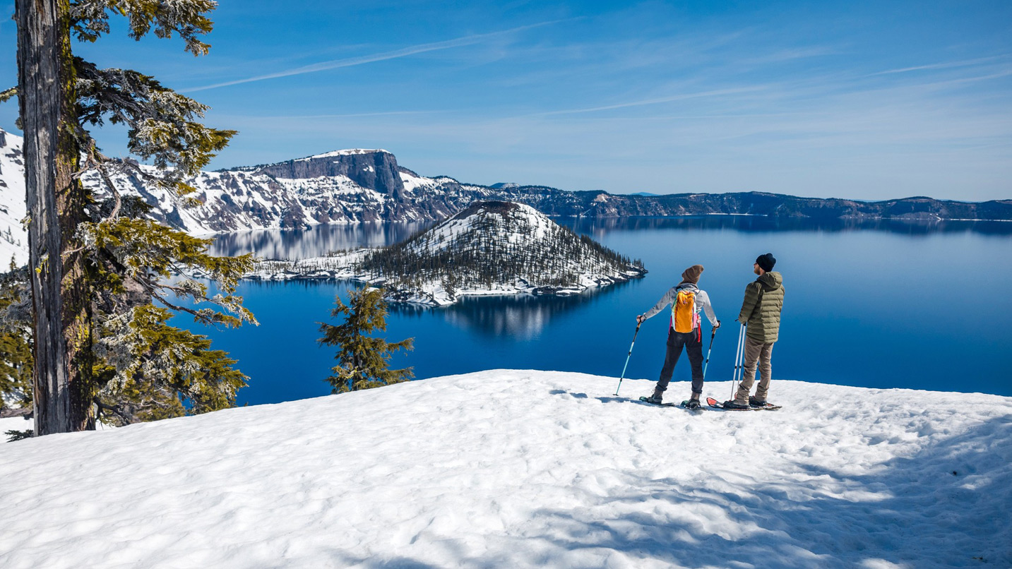 Two people in snowshoes stand on a snowy overlook, gazing at Crater Lake's Wizard Island, surrounded by rugged, snowy cliffs under a clear blue sky.