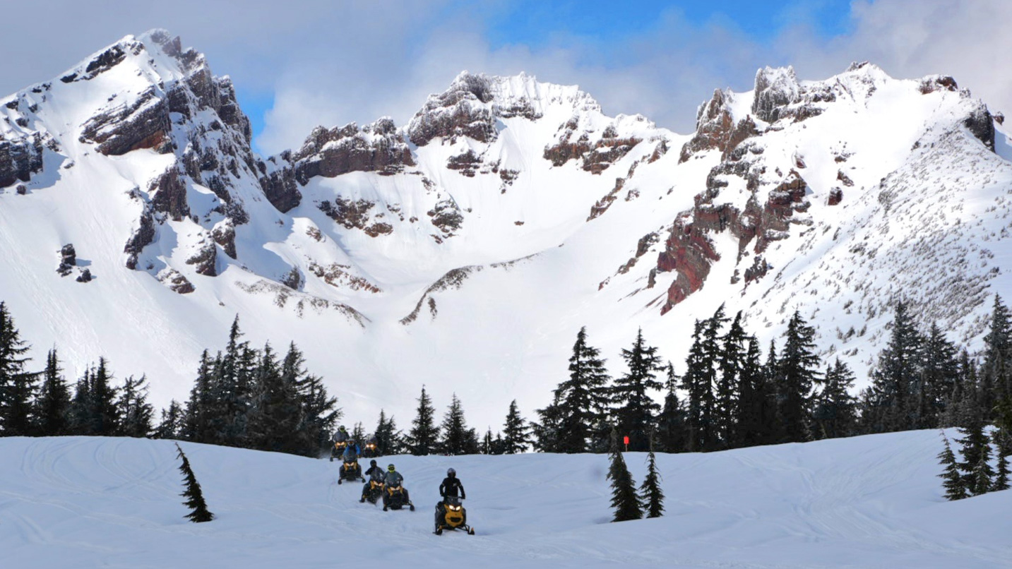 A group of snowmobilers in a single line traverse a snowy expanse with epic views of a snow-capped mountain range in the background.