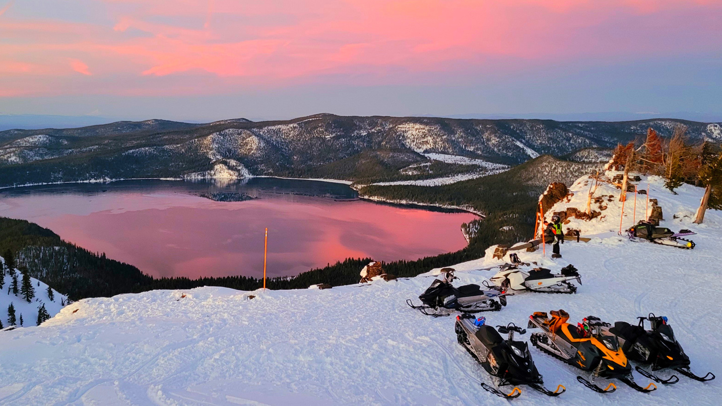 Snowmobiles parked near a snowy viewpoint of a large lake.
