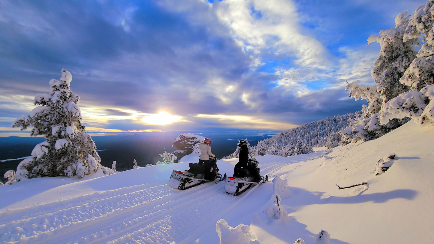 Two snowmobile riders pause on a snowy track to enjoy a sunset.