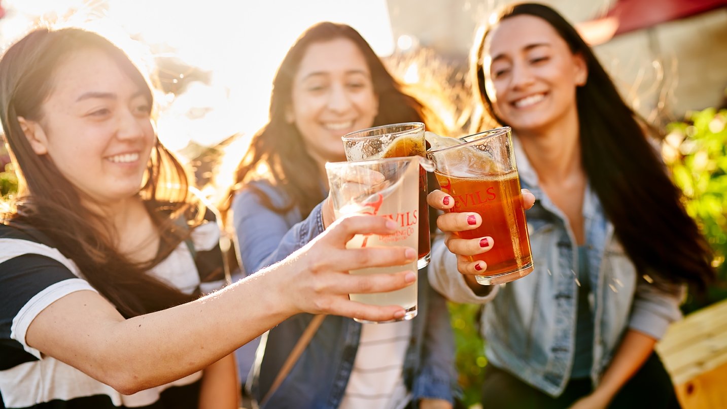 Three women with black hair hold pint glasses of beer in a cheers toast, with sunlight behind them.