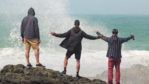 Three people stand on rocks with arms outstretched facing the ocean