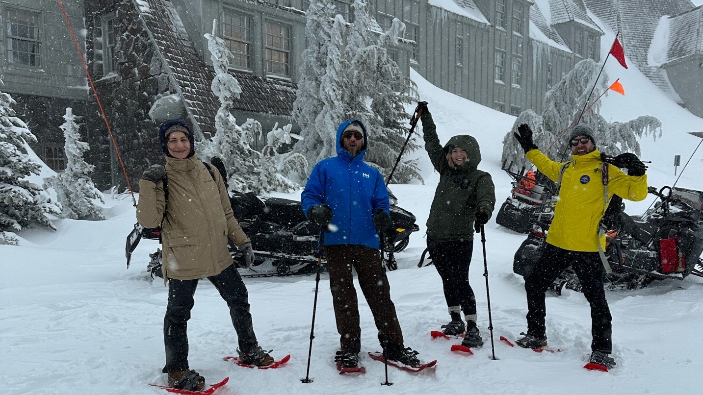 Four people wear coats and snowshoes in snow with large building and snowmobiles in background.