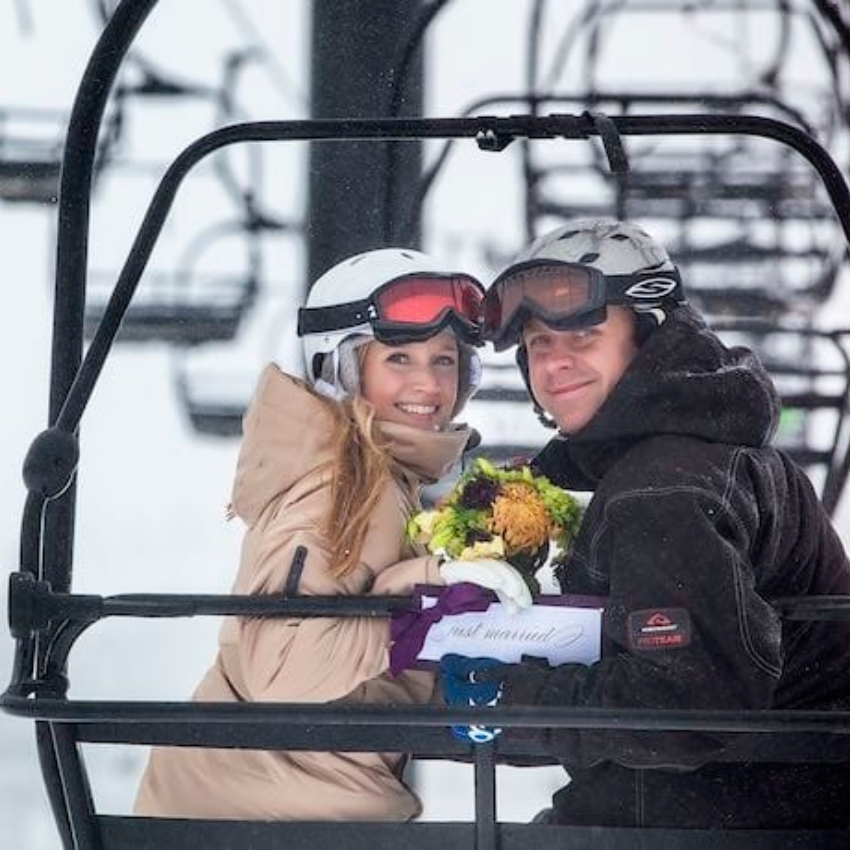 Man and woman with ski goggles, helmet and coats smile while seated on chairlift.