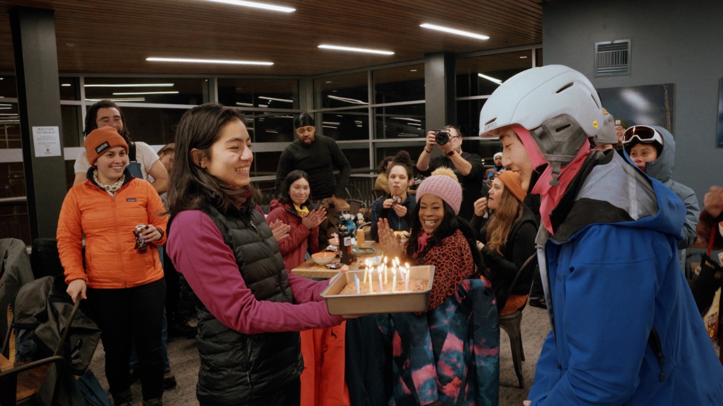 Person prepares to blow out birthday candles as a room full of people in winter gear smile in background