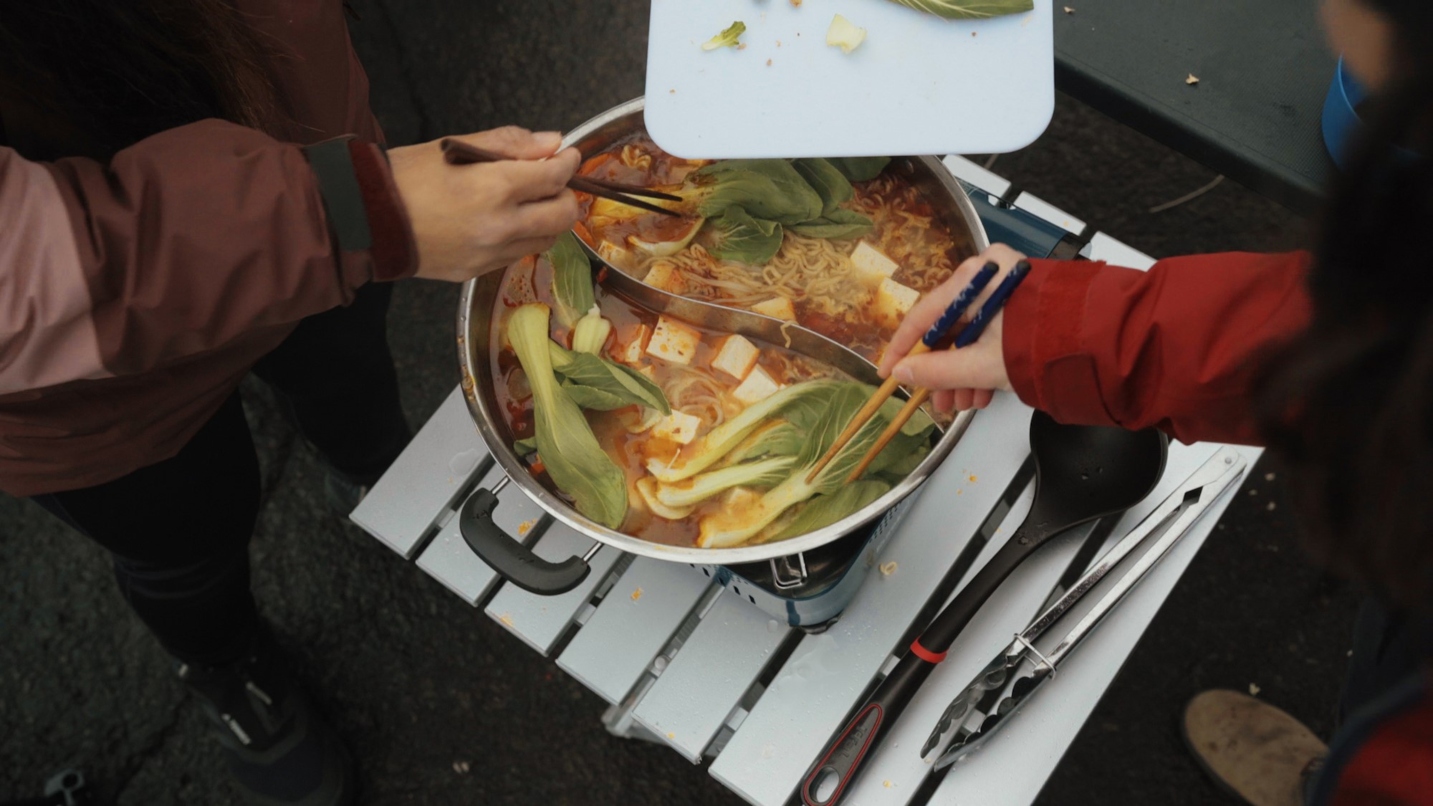 Two people reach chopsticks into pot of noodles, broth and greens on a portable table