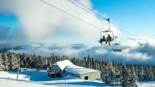 Two people sit on ski lift against field of snow and blue sky