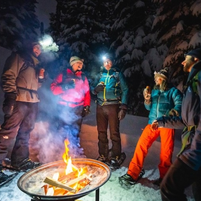 A group gathers around a bonfire in the snow at night.