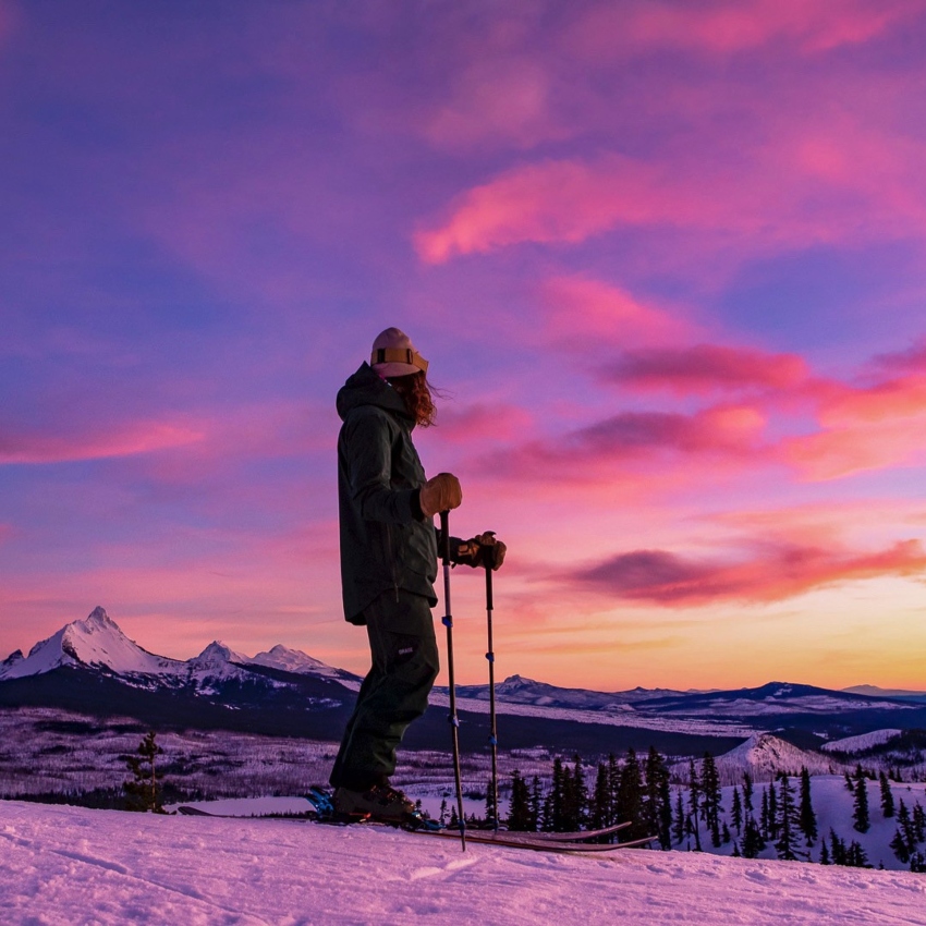 A single skier looks out over a purple and pink sunset.