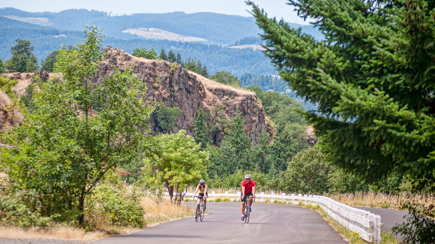 Two people biking on a road set against views and foliage of the Gorge.