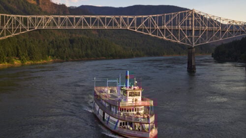 A vintage style large cruise boat passes underneath a bridge spanning over the Columbia River.