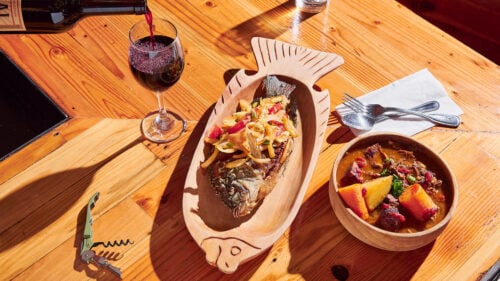 A roasted whole fish with vegetables on a wooden platter shaped as a fish, next to a bowl of meat stew and a glass of wine.
