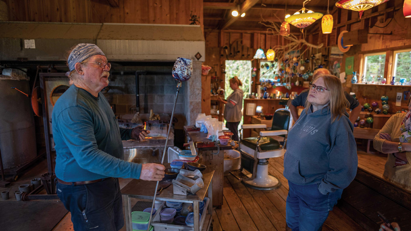 A glassblower demonstrating a piece still on the punty to a woman.