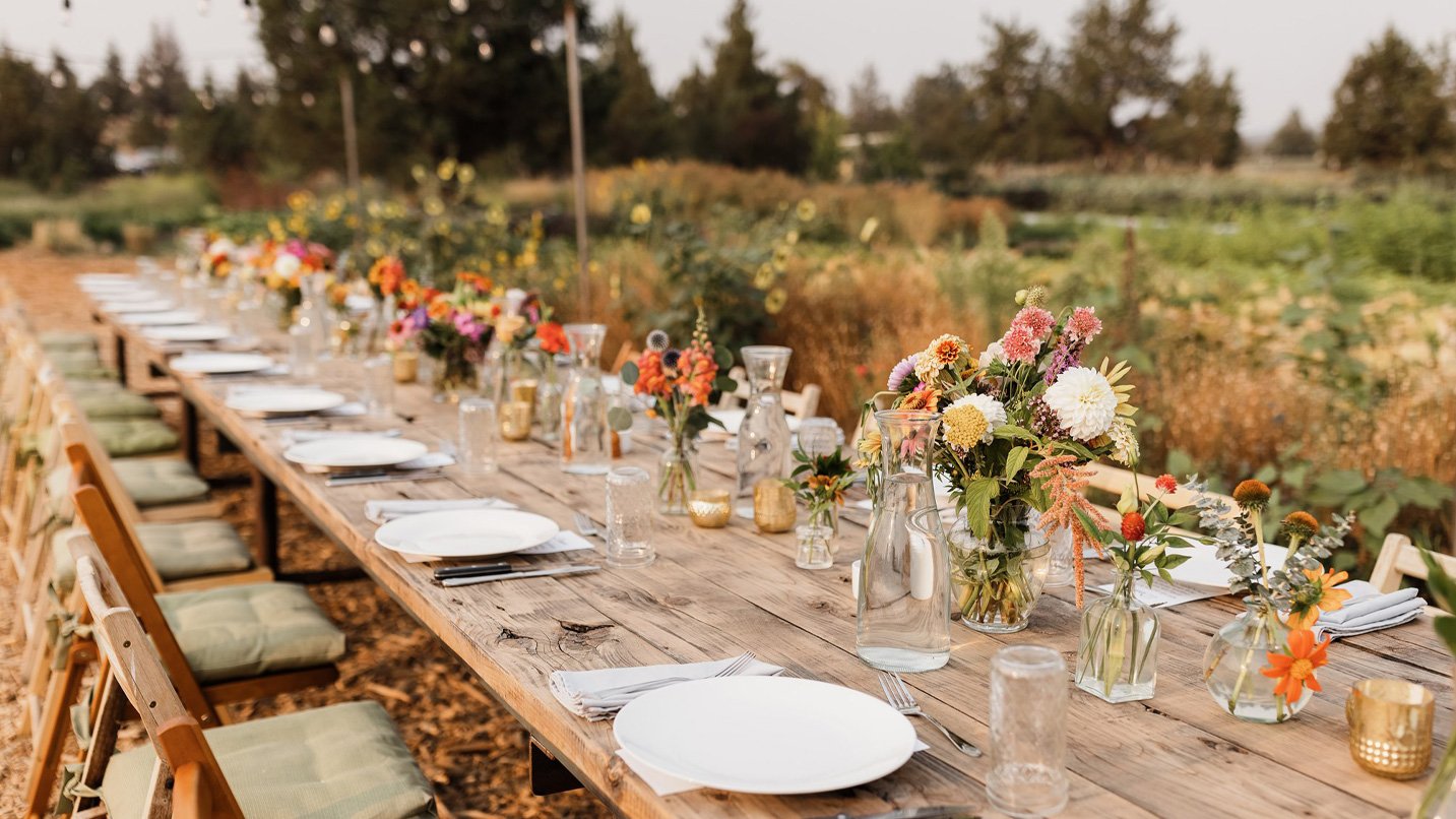 A long wooden table is set with bouquets of flowers in a farm field.
