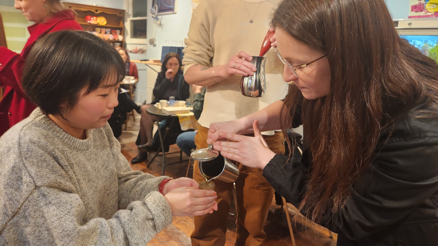 Woman with short black hair holds small cup as woman with long brown hair pours a liquid from metal vessel into the cup