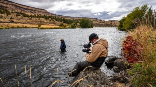 Man sits on rock holding camera pointed at man wading in river