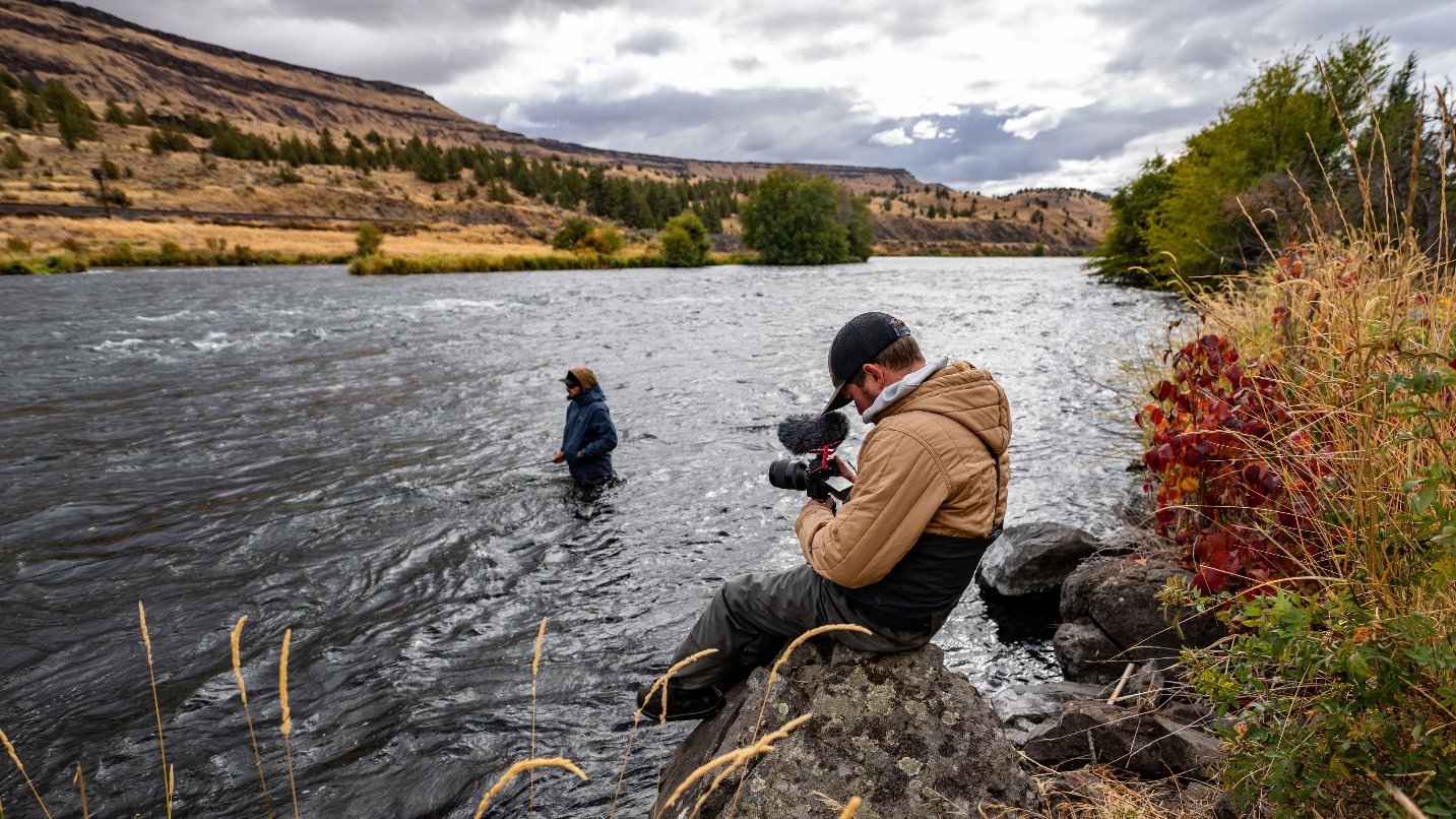 Man sits on rock holding camera pointed at man wading in river
