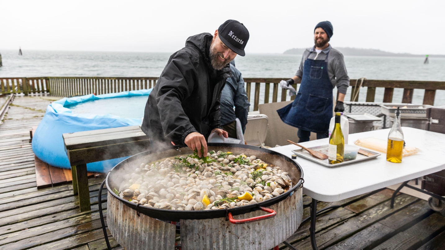 Man puts green ingredient on large outdoor cooking vessel of shellfish, on a dock on the ocean