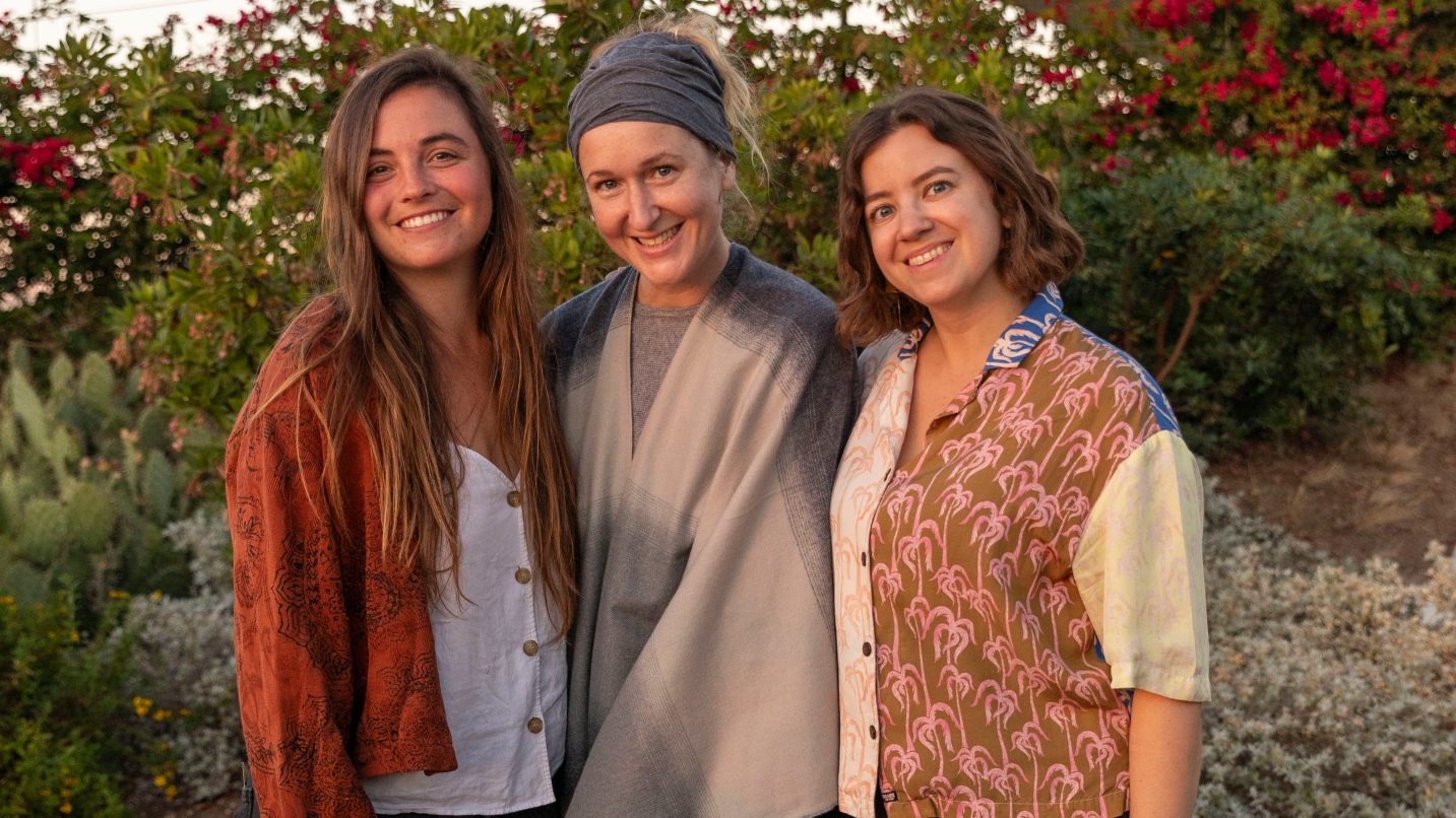 three women smile at camera with row of bushes and red flowers in background