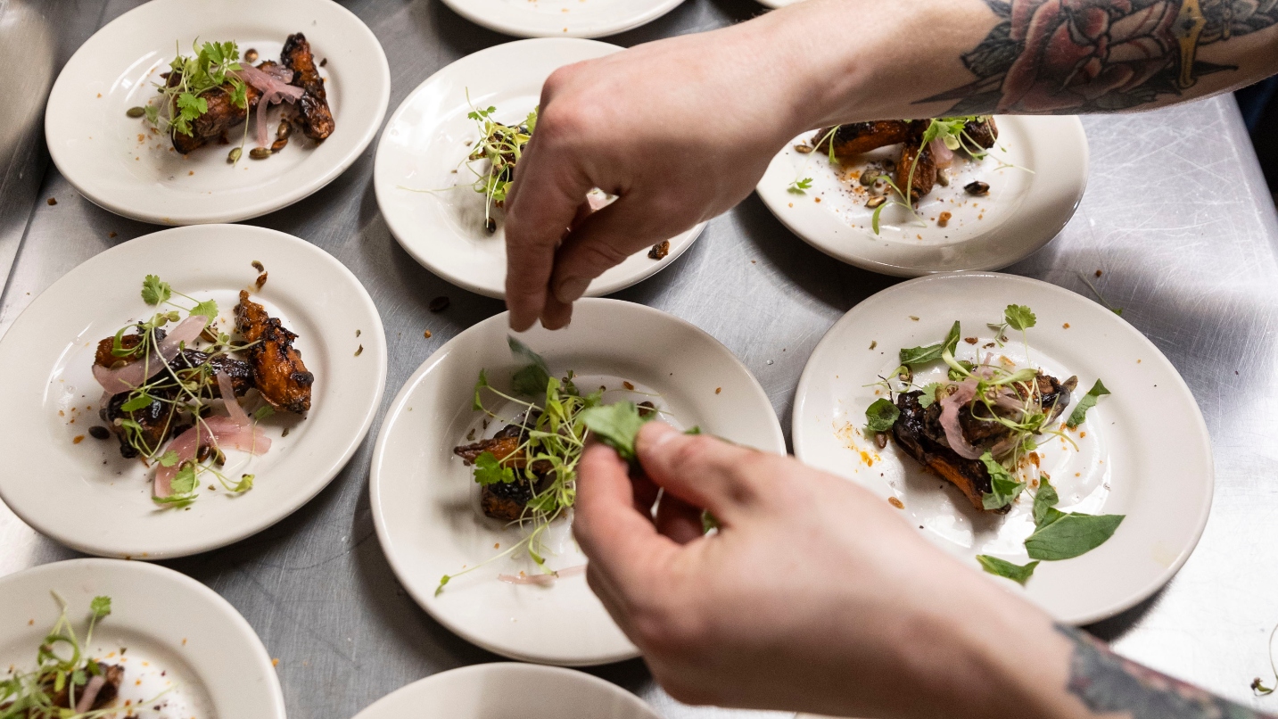 Two arms with tattoos add green herbs to small plates of food