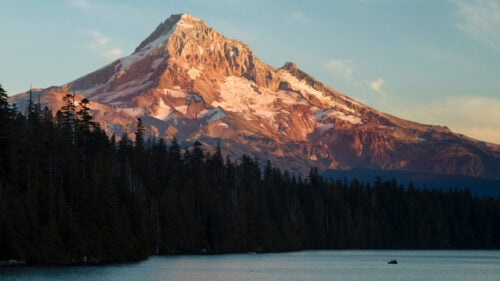 Landscape of Mt. Hood during sunset, with large conifers and a lake with a small boat in the foreground.