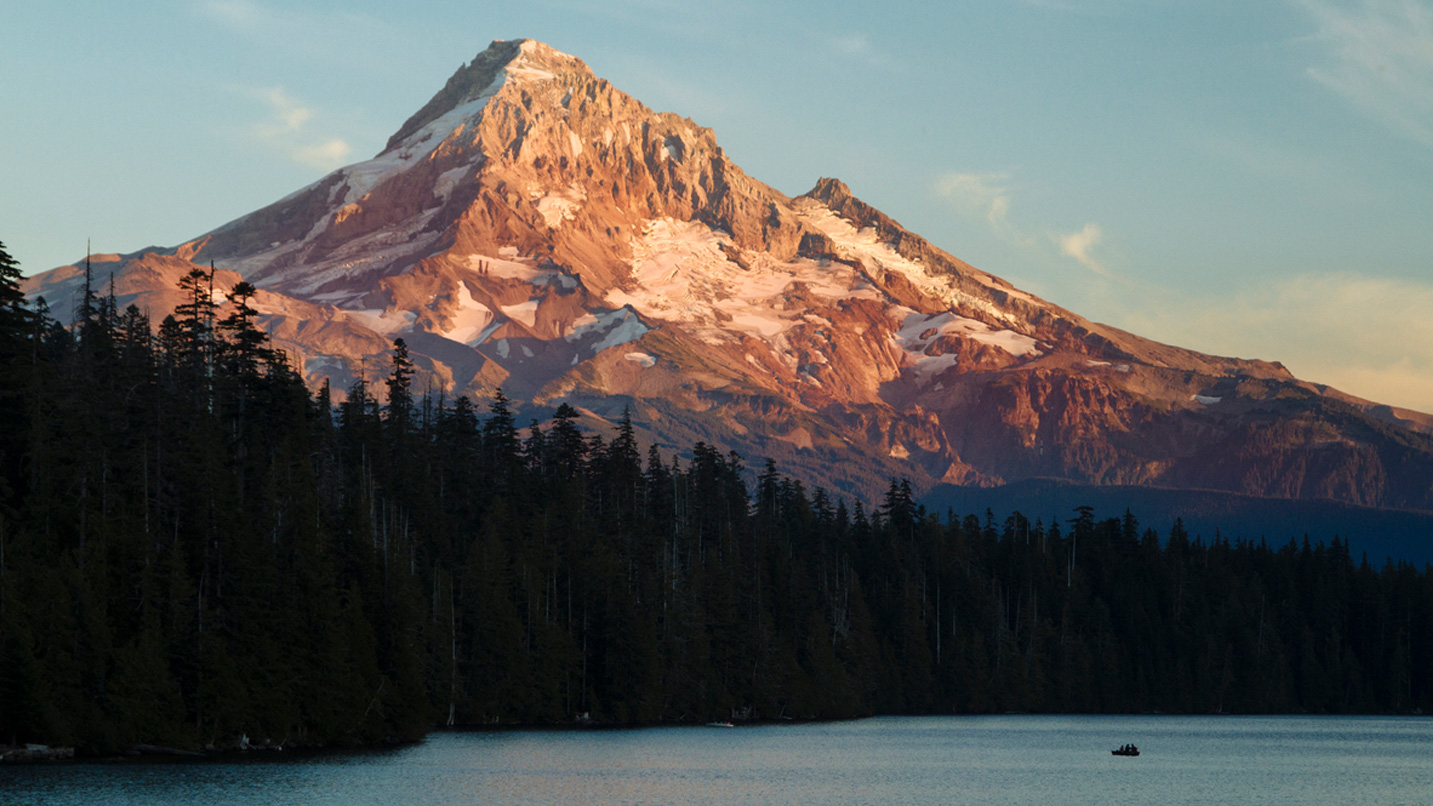 Landscape of Mt. Hood during sunset, with large conifers and a lake with a small boat in the foreground.