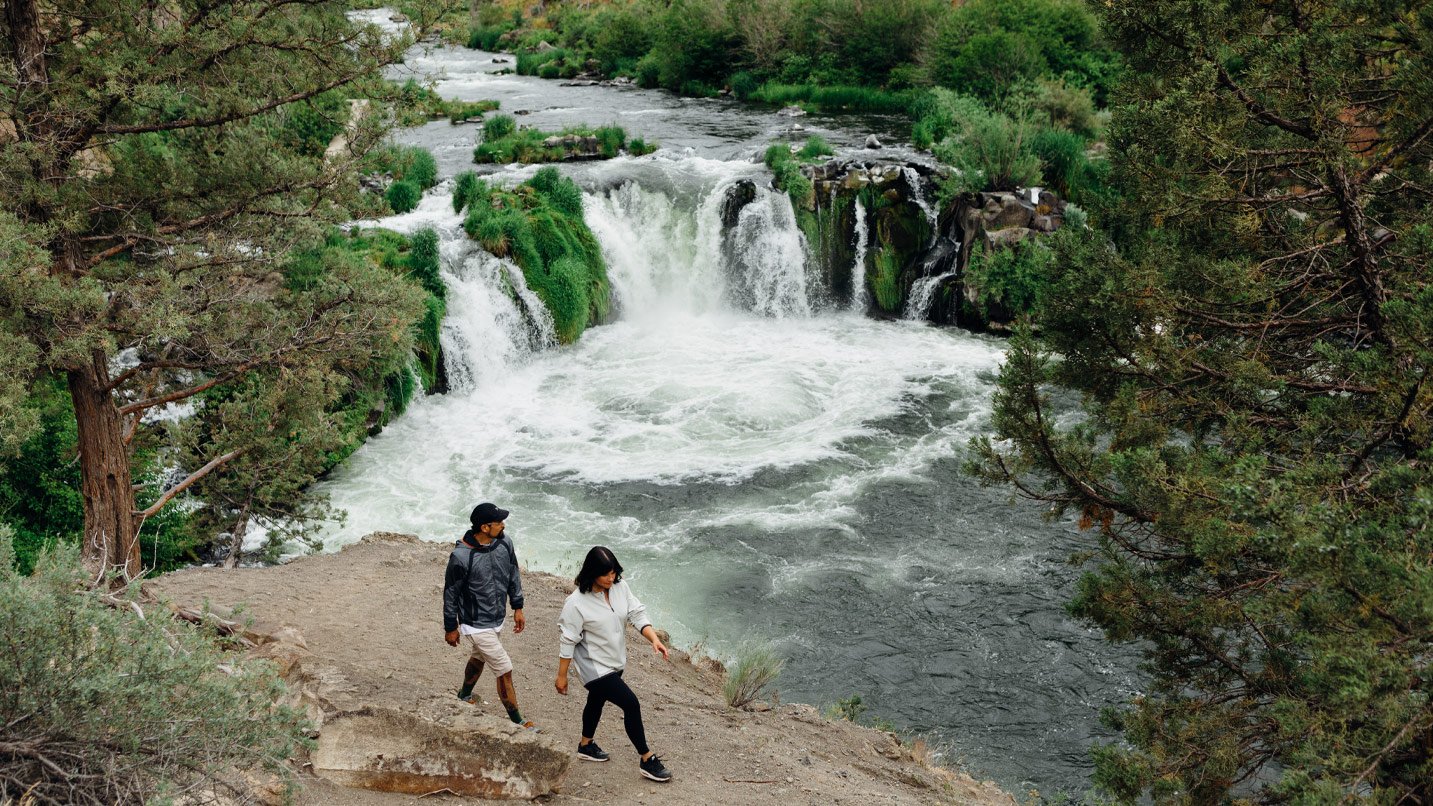 Two people hike along a trail next to a waterfall.