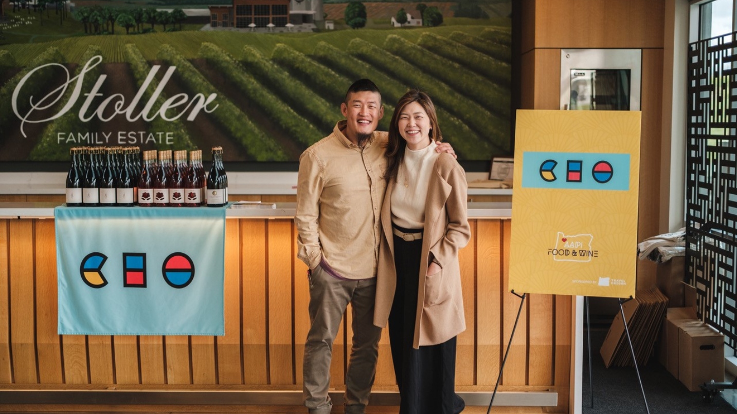 Man and woman stand in front of winery signage and counter with wine bottles