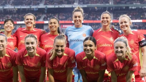 Two rows of women wear athletic shirts and smile at the camera with stadium in background