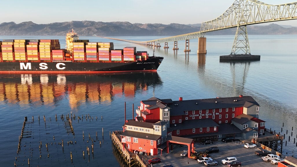 A hotel on a dock has views of a cargo ship traveling under a bridge.