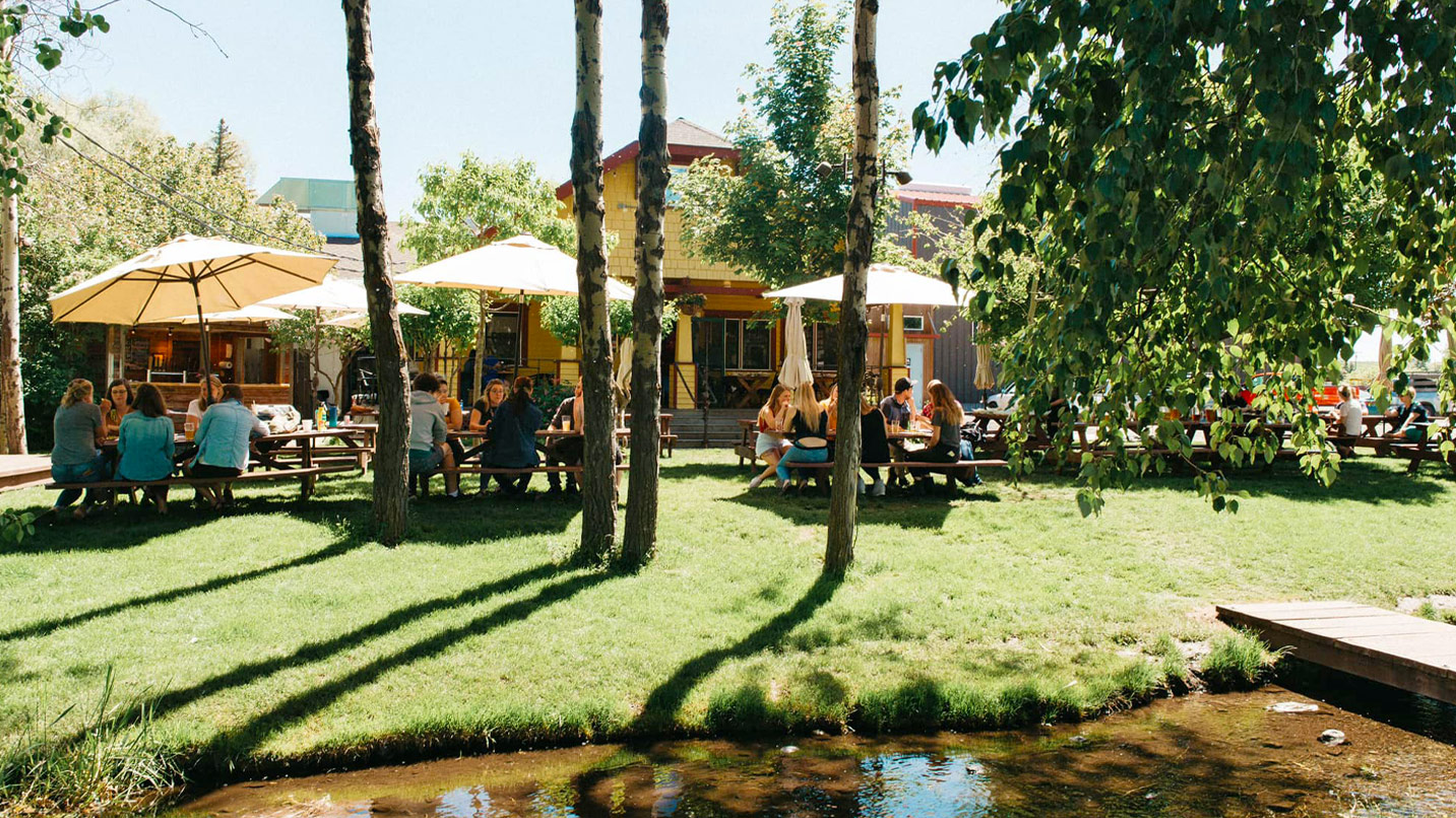 Several groups at shaded picnic tables outdoors behind a restaurant near a creek.