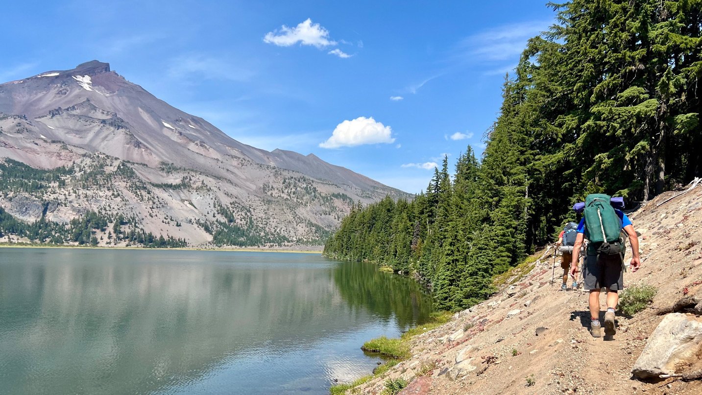 A backpacker hikes along a lake at the base of a mountain.