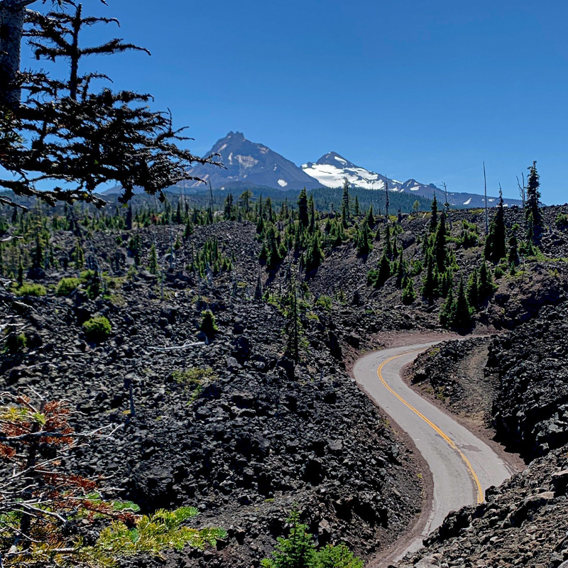 A road curves through a lava field with snow-capped peaks in the distance.